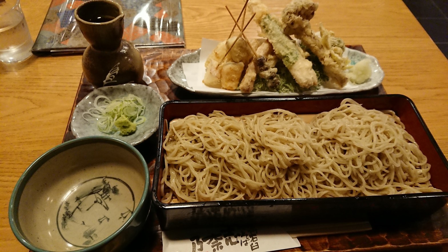 黒塗り角ざるに盛られた蕎麦、陶器の蕎麦猪口、奥に天ぷらを積み上げた皿と徳利